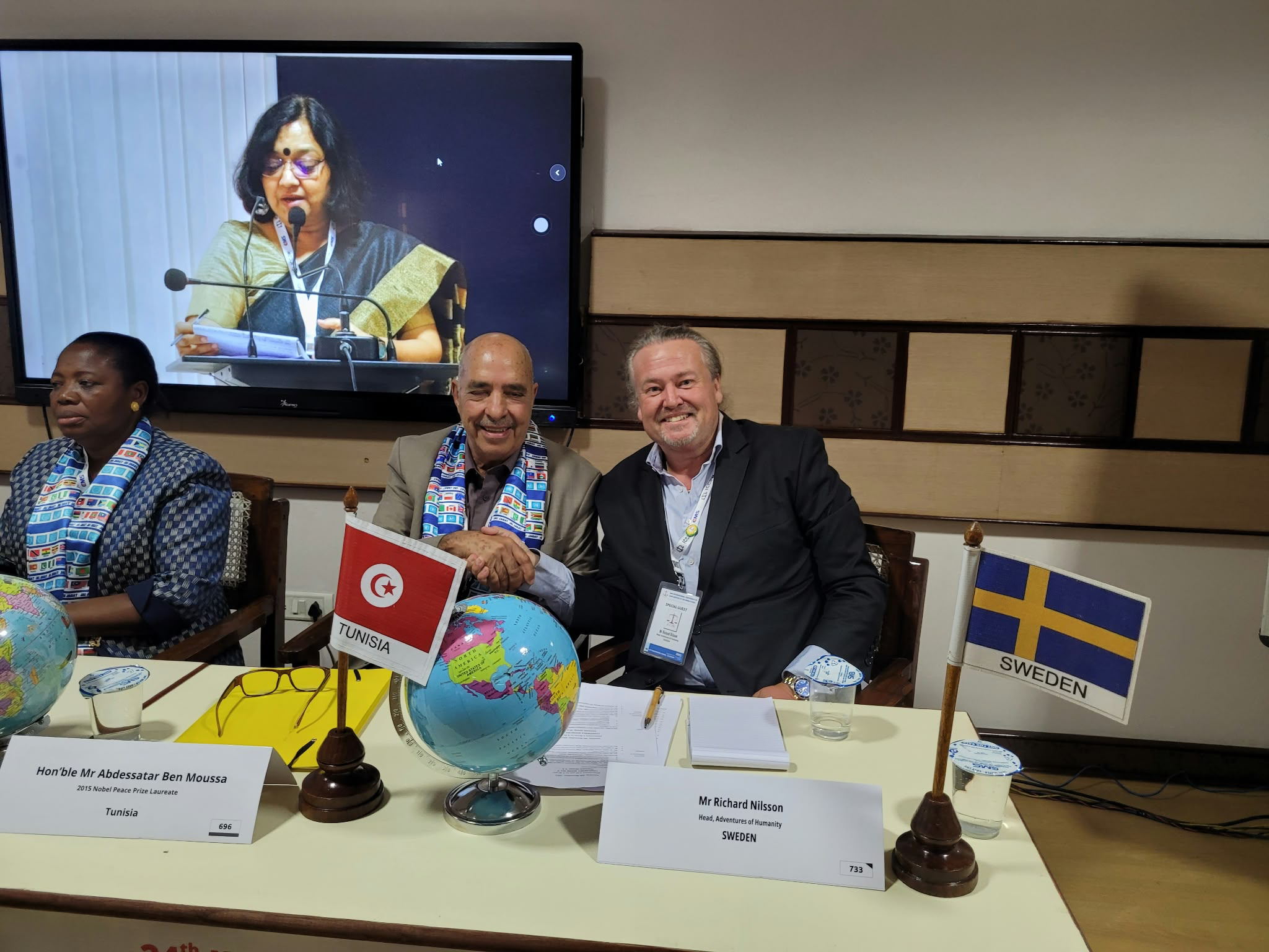 Tunisia और Sweden के झंडों वाली मेज़ के पास अंतरराष्ट्रीय बैठक में एक प्रतिनिधि से हाथ मिलाते Richard Nilsson